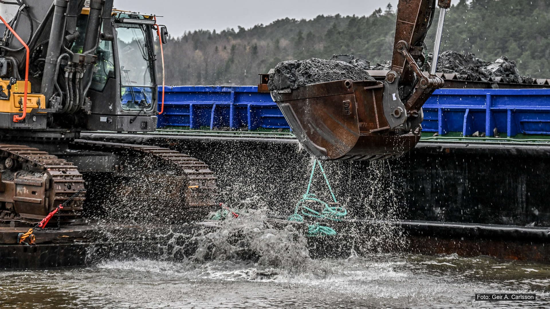 Gravemaskin på lekter mudrer sjøbunn i havneområde, med vannsprut og masser som løftes opp.